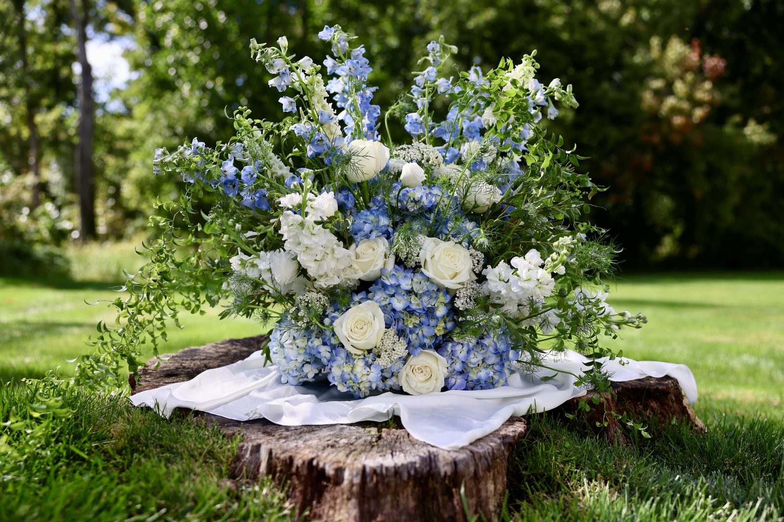 A large arrangement of blue delphinium, hydrangea and white roses on a tree stump draped in white fabric