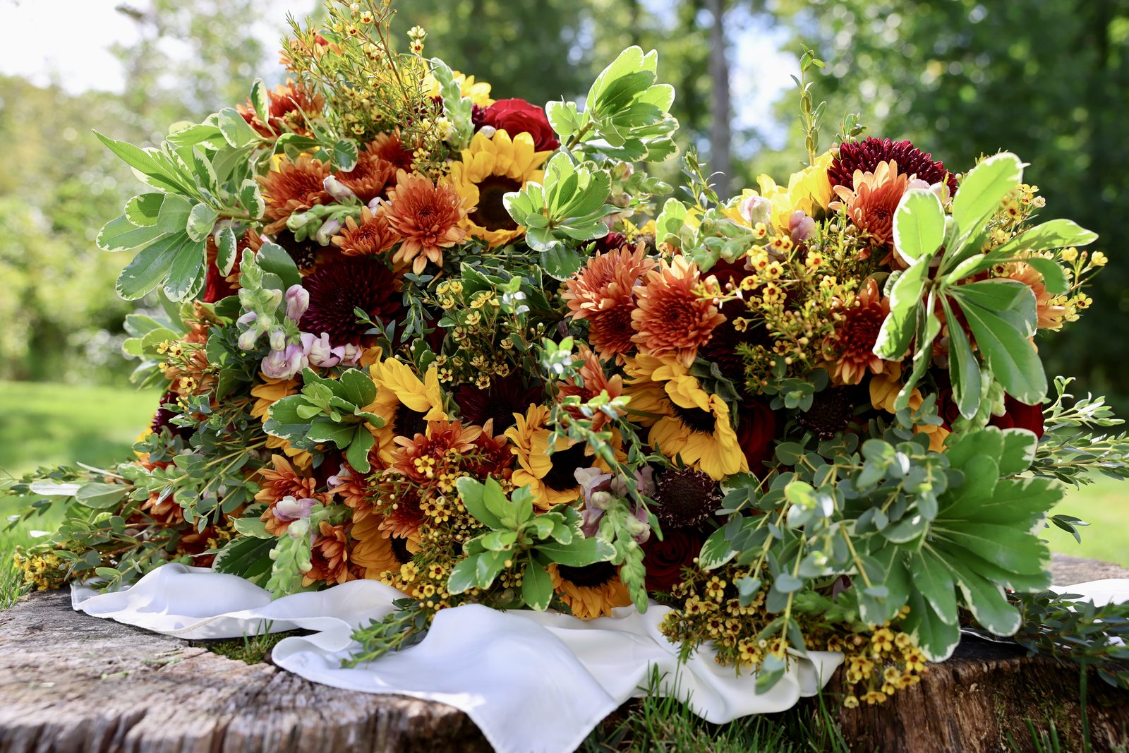 An autumn arrangement of sunflowers, rust chrysanthemums, red roses and waxflower on a tree stump